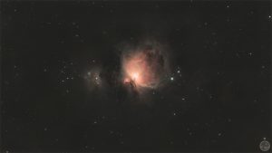 Fotografia astronómica de campo amplo da Grande Nebulosa de Orionte (M42), exibindo a estrutura nebulosa rosada e circular concentrada no centro do plano, com o núcleo brilhante bem definido e a nebulosa vizinha M43, rodeadas por um vasto campo pontilhado de estrelas de fundo sobre o éter negro.