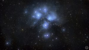 Fotografia astronómica de campo amplo do aglomerado estelar das Plêiades (M45), exibindo um grupo central de estrelas azuis brilhantes envolto numa intensa e detalhada nebulosidade filamentosa de cor azul-clara, rodeada por inúmeras estrelas de fundo mais pequenas sobre o éter negro profunda.