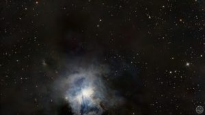 Fotografia astronómica profunda exibindo a Nebulosa da Íris (NGC 7023) no centro, caracterizada por um núcleo estelar brilhante intensamente rodeado por delicadas e complexas nuvens de poeira reflectora de tons azuis e anil. Todo o campo em torno da nebulosa revela extensas e imponentes massas de poeira escura e acastanhada que bloqueiam a luz de fundo. A imagem encontra-se ponteada por estrelas nítidas de diversas colorações sobre um céu negro e profundo.