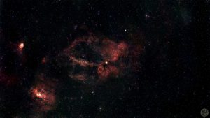 Fotografia astronómica de campo amplo destacando um enorme anel incompleto de gás vermelho rubro com a forma de uma garra, correspondente à nebulosa SH2-157. A estrutura gasosa é complexa, densa e esculpida, destacando-se veios mais escuros de poeira. No quadrante inferior esquerdo brilha uma nebulosa secundária bastante compacta e intensa (NGC 7538). O fundo negro do espaço é imaculado, salpicado por estrelas pontuais perfeitas cujas cores individuais se mantêm bem preservadas.