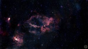 Fotografia astronómica de campo amplo destacando um enorme anel incompleto de gás vermelho rubro com a forma de uma garra, correspondente à nebulosa SH2-157. A estrutura gasosa é complexa, densa e esculpida, destacando-se veios mais escuros de poeira. No quadrante inferior esquerdo brilha uma nebulosa secundária bastante compacta e intensa (NGC 7538). O fundo negro do espaço é imaculado, salpicado por estrelas pontuais perfeitas cujas cores individuais se mantêm bem preservadas.