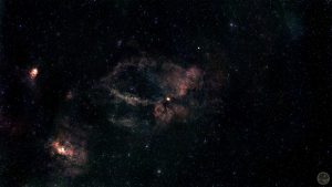 Fotografia astronómica de campo amplo destacando um enorme anel incompleto de gás vermelho rubro com a forma de uma garra, correspondente à nebulosa SH2-157. A estrutura gasosa é complexa, densa e esculpida, destacando-se veios mais escuros de poeira. No quadrante inferior esquerdo brilha uma nebulosa secundária bastante compacta e intensa (NGC 7538). O fundo negro do espaço é imaculado, salpicado por estrelas pontuais perfeitas cujas cores individuais se mantêm bem preservadas.