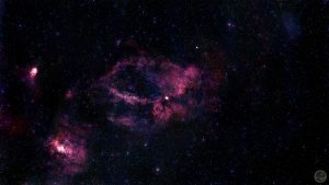 Fotografia astronómica de campo amplo destacando um enorme anel incompleto de gás vermelho rubro com a forma de uma garra, correspondente à nebulosa SH2-157. A estrutura gasosa é complexa, densa e esculpida, destacando-se veios mais escuros de poeira. No quadrante inferior esquerdo brilha uma nebulosa secundária bastante compacta e intensa (NGC 7538). O fundo negro do espaço é imaculado, salpicado por estrelas pontuais perfeitas cujas cores individuais se mantêm bem preservadas.